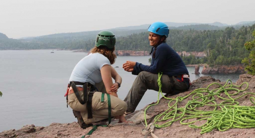 Two people wearing safety gear are secured by ropes near the edge of a cliff. One person appears to be an instructor, giving direction to the other person. There is a lake below them.